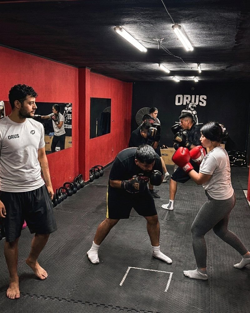Alumnos practicando boxeo con guantes en una clase de ORUS, con un instructor mirando a la izquierda, frente a la pared roja del gimnasio